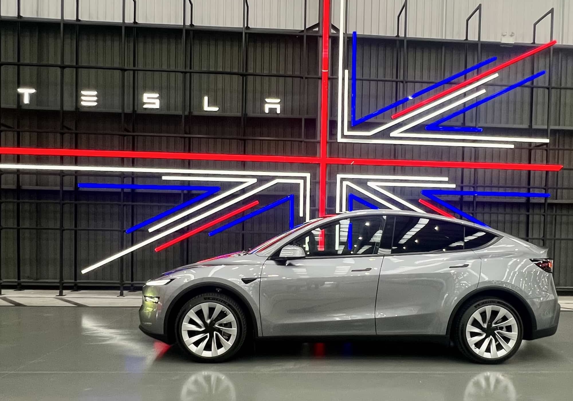 A silver2025 tesla Model Y is parked in front of a neon union jack and tesla sign ready to be collected from teslas Longbridge collection point